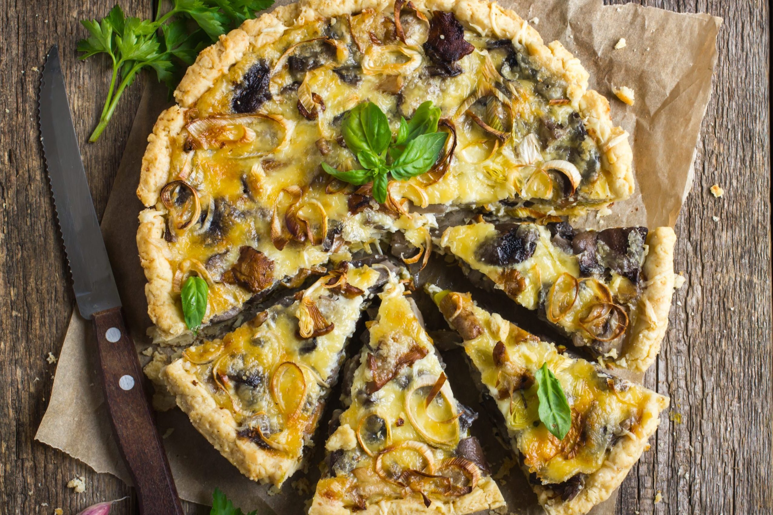 Quiche with mushrooms and onions, partially sliced, on parchment paper, surrounded by parsley on a rustic wooden table, with a serrated knife nearby.