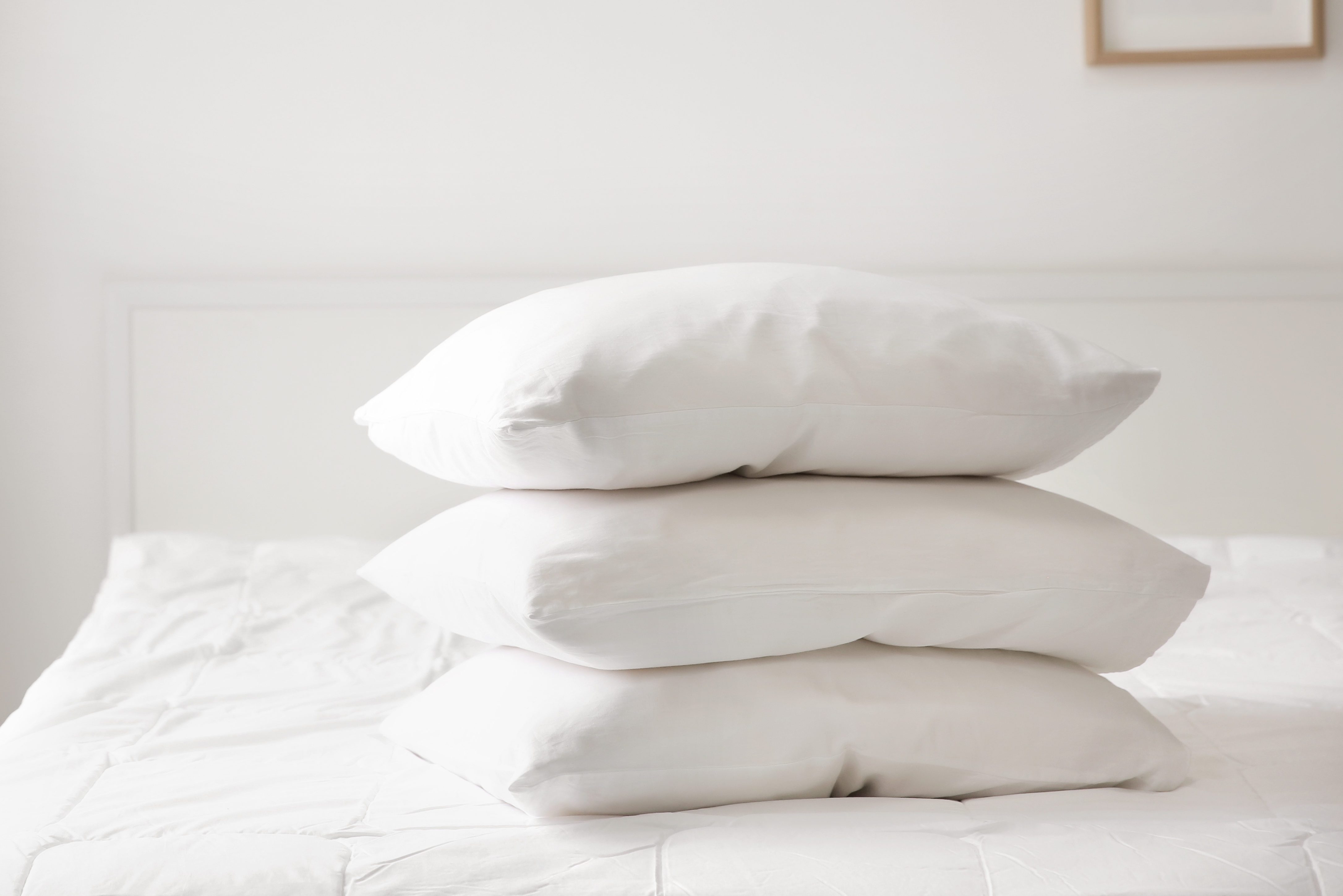 Three white pillows stacked neatly on a white bedspread in a minimalistic, bright room.