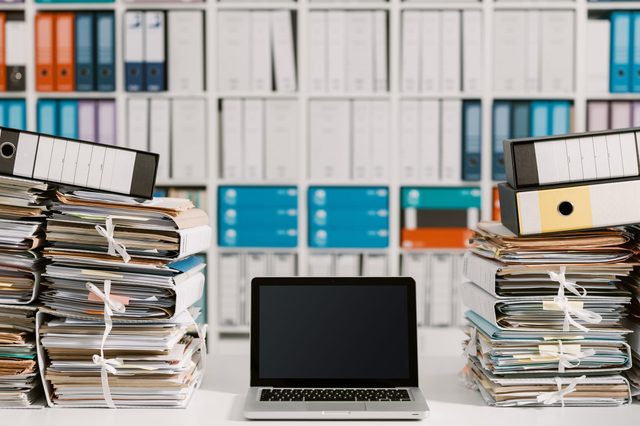A laptop sits on a desk surrounded by organized stacks of paperwork and binders, with a backdrop of neatly arranged shelves.