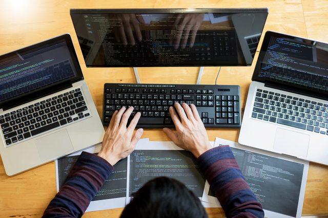 A person types code on a keyboard surrounded by two laptops and code-printed papers, placed on a wooden desk.
