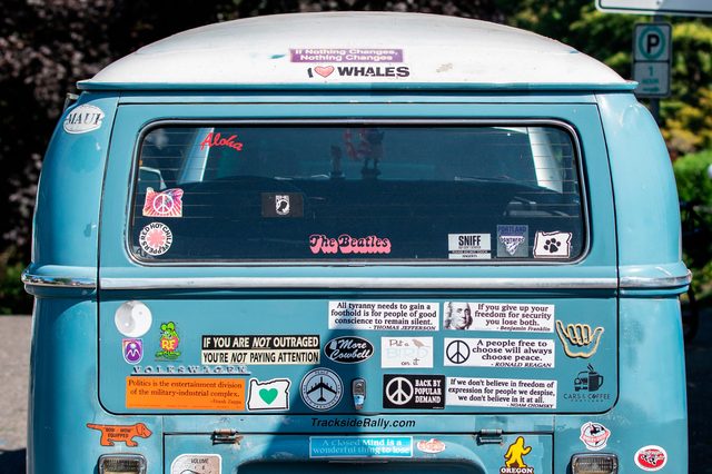 A blue van displays numerous bumper stickers with messages about peace and freedom, surrounded by trees and a parking sign.