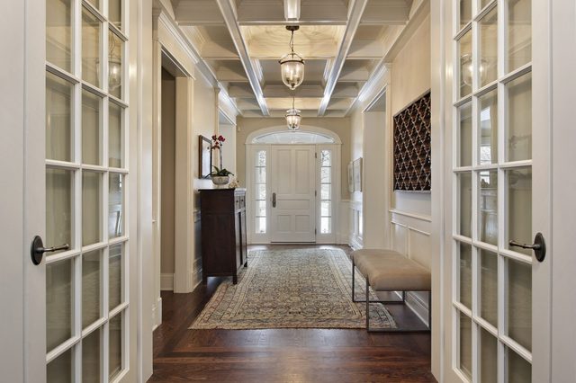 A hallway features a patterned rug leading to a front door, with glass side panels, ornate ceiling, and elegant furnishings on hardwood floor.