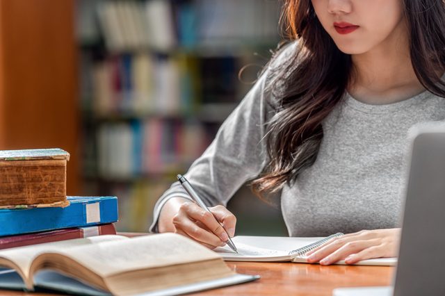 Woman writing notes in a notebook, surrounded by open books, in a library setting.