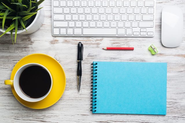 Keyboard, mouse, blue notebook, and pencil rest on a wooden desk beside a yellow saucer with coffee and a green plant.