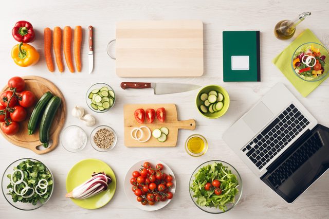 Vegetables sit beside a laptop on a kitchen countertop with cutting boards, knives, oil, and a notebook, creating a cooking preparation scene.