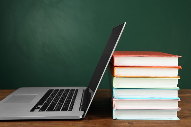 Laptop leans against stacked books on a wooden table, with a green chalkboard background.