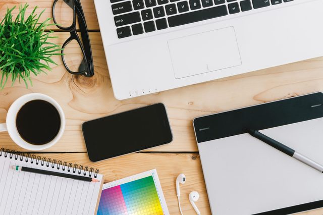 Laptop rests on wooden desk, surrounded by glasses, coffee cup, smartphone, notebook, color chart, earphones, and drawing tablet with stylus.
