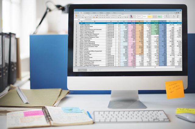 A computer displays a spreadsheet with financial data beside scattered notebooks and sticky notes in a bright office environment.