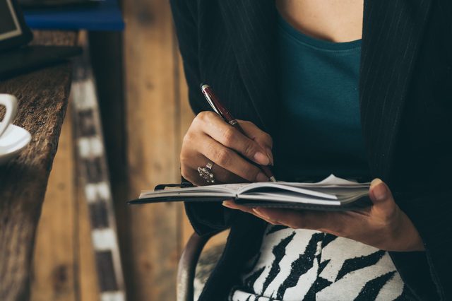 Person writing in a notebook while seated at a wooden table, next to a white coffee cup, in a cozy, rustic setting.