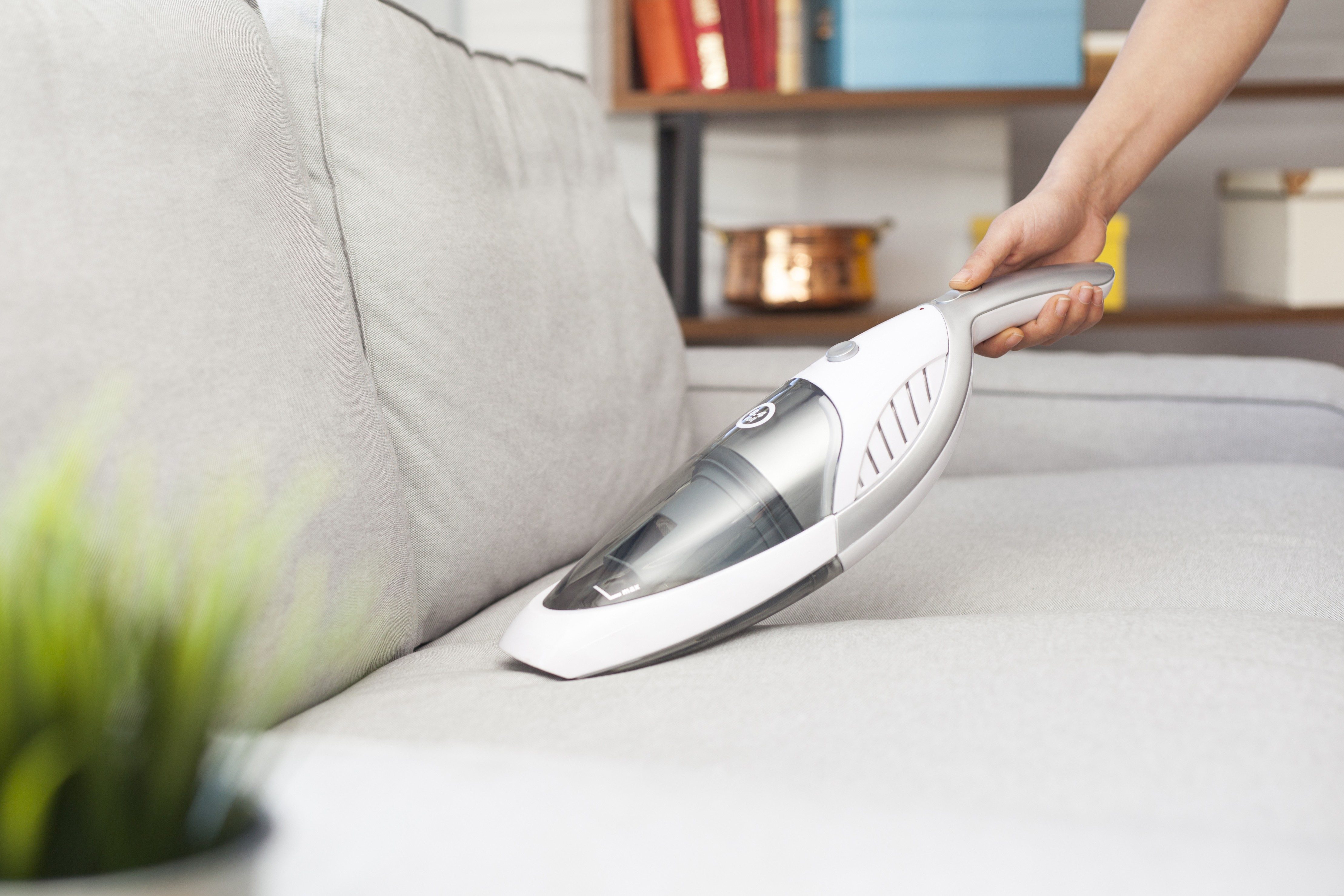 Handheld vacuum cleans gray sofa; shelf with books and copper pot in the background.