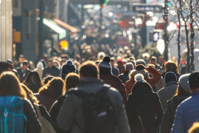 Crowd walking on a busy city street, wearing winter clothing, surrounded by buildings and storefronts.