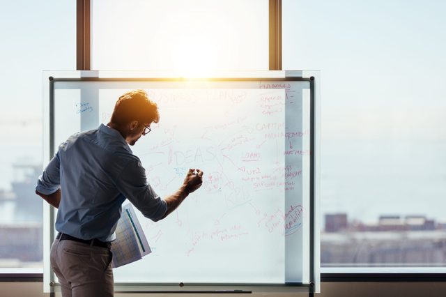 Person writing on a whiteboard covered in ideas and diagrams, holding a paper, with a sunlit cityscape visible through the window.