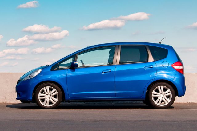 A blue car is parked on a paved surface against a concrete barrier, with a blue sky and scattered clouds above.