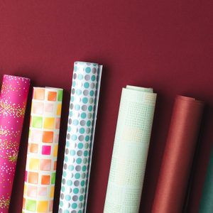Colorful rolls of wrapping paper stand upright on a red background, featuring various patterns like polka dots, squares, and chevrons.