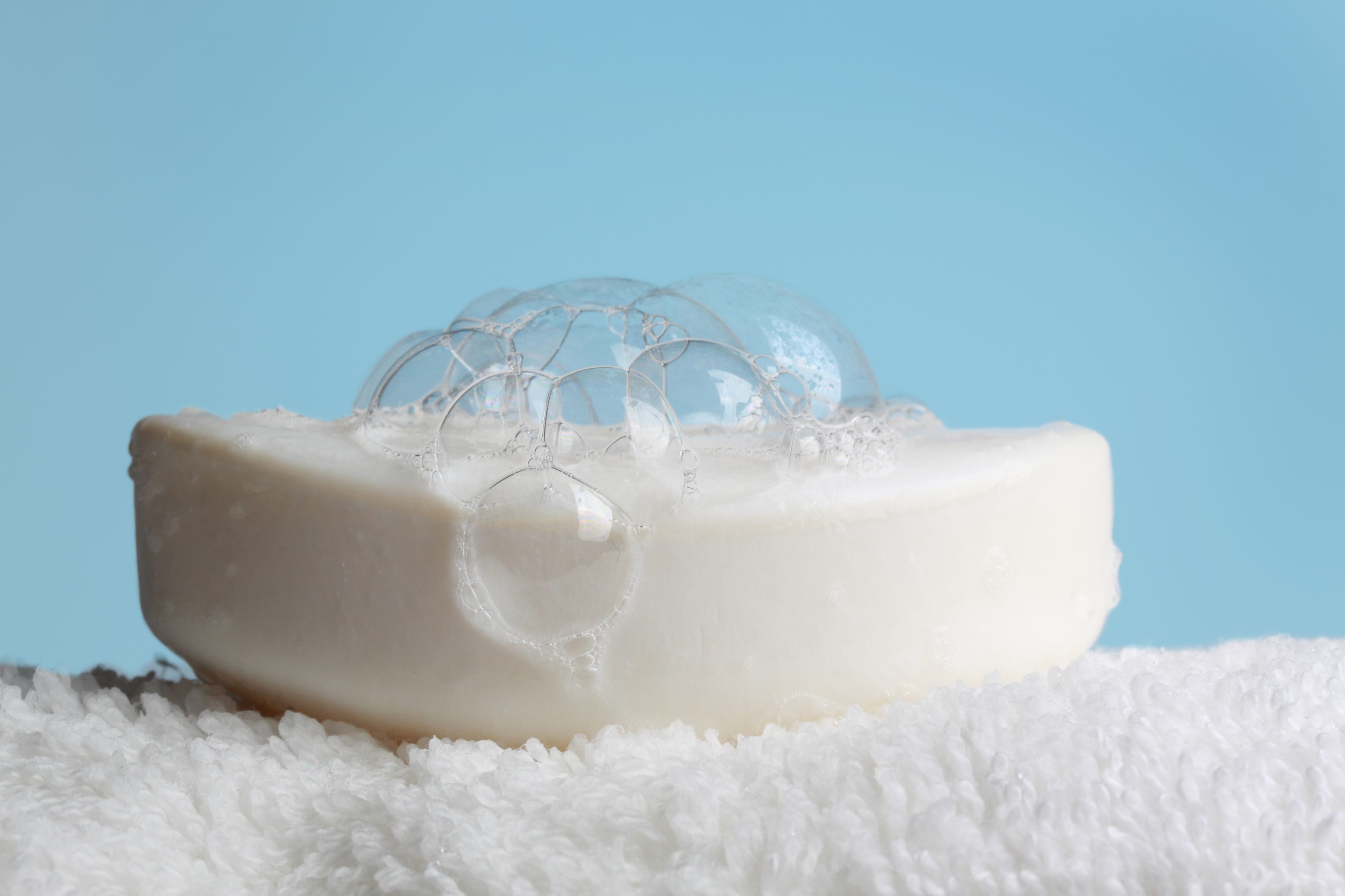 Soap bar with bubbles sits on a fluffy, white towel against a light blue background.
