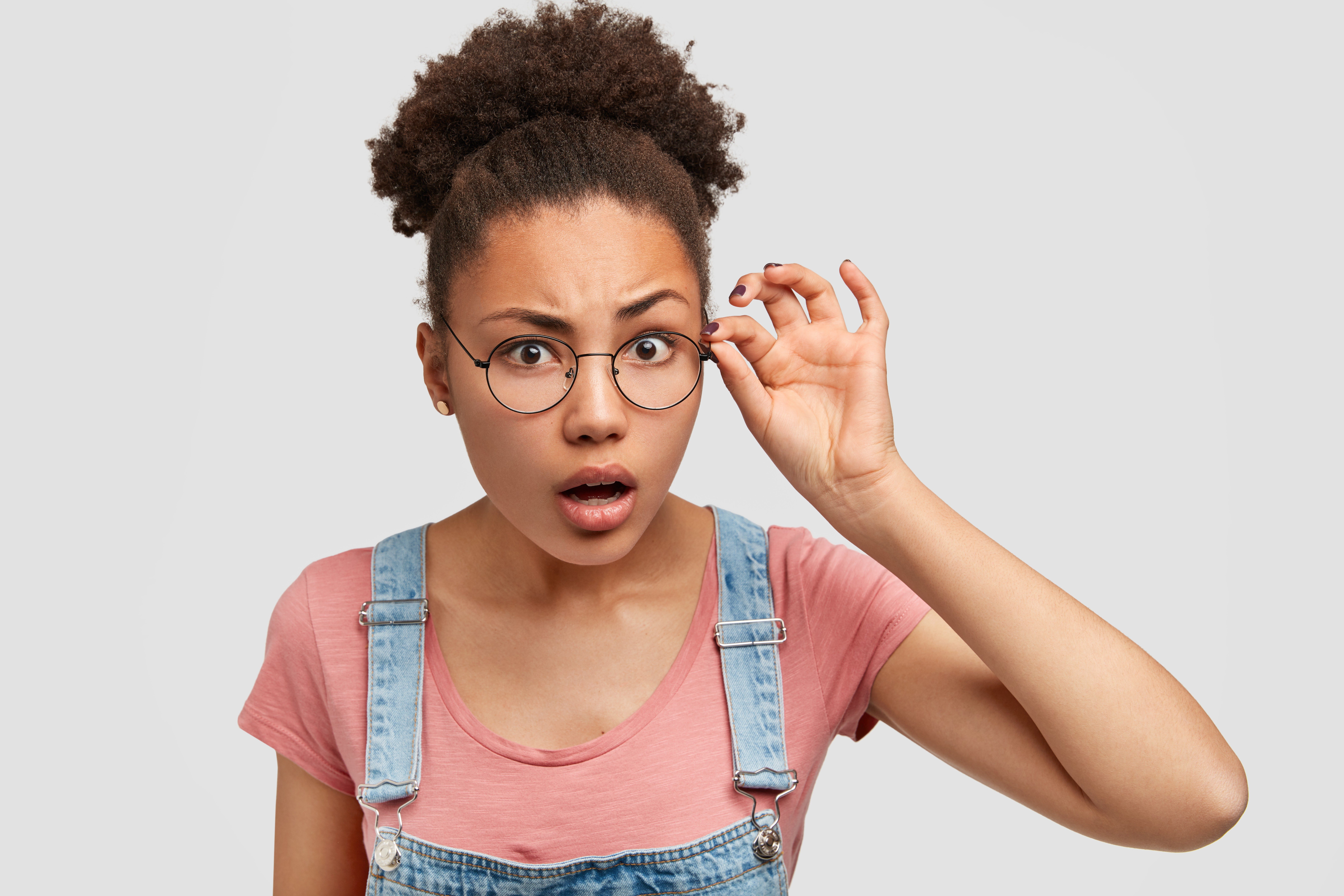 Person adjusts round glasses, expression surprised, wearing pink shirt and denim overalls, against plain gray background.
