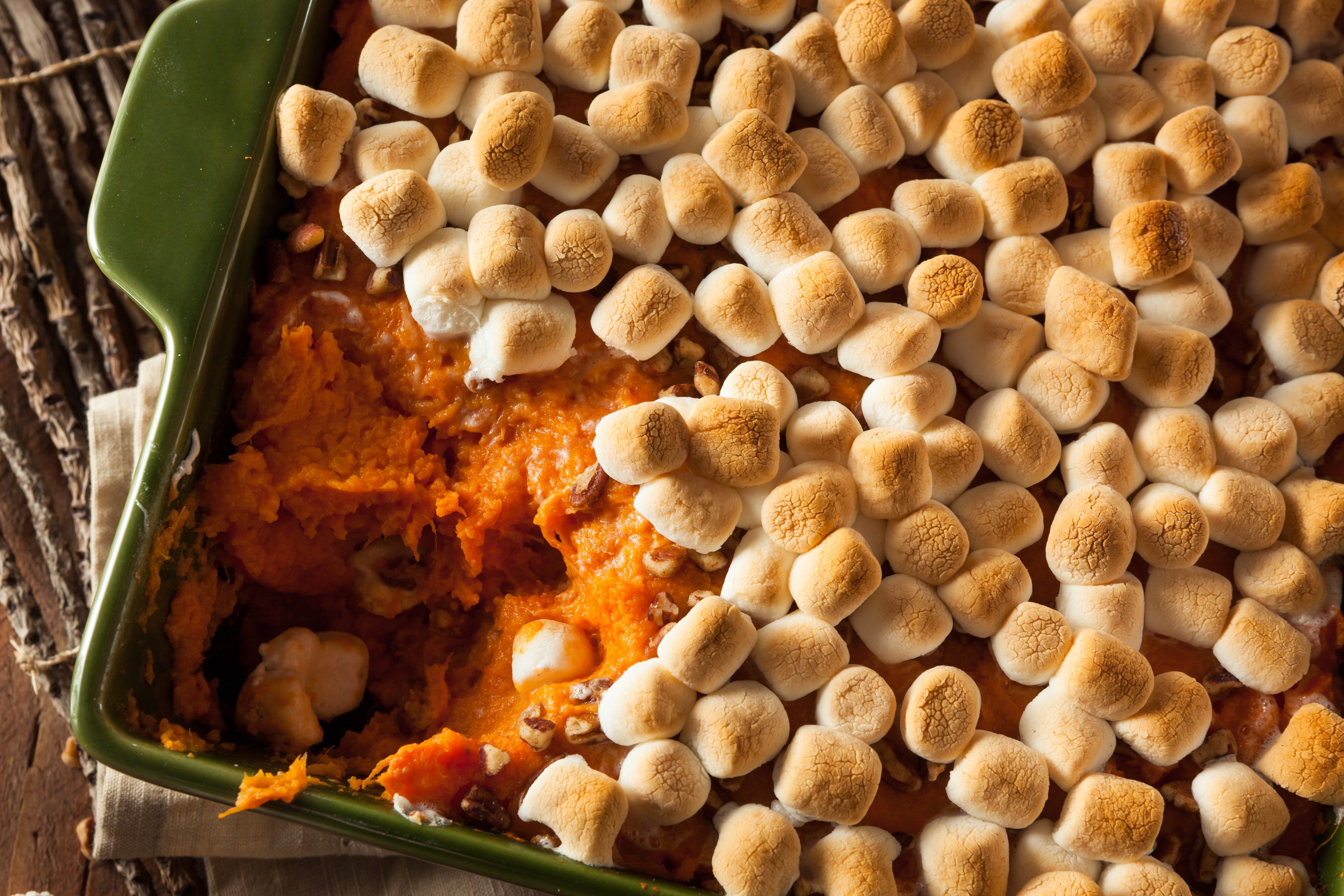 Marshmallow-topped sweet potato casserole, partially eaten, sits in a green baking dish on a wooden table with rustic decor.