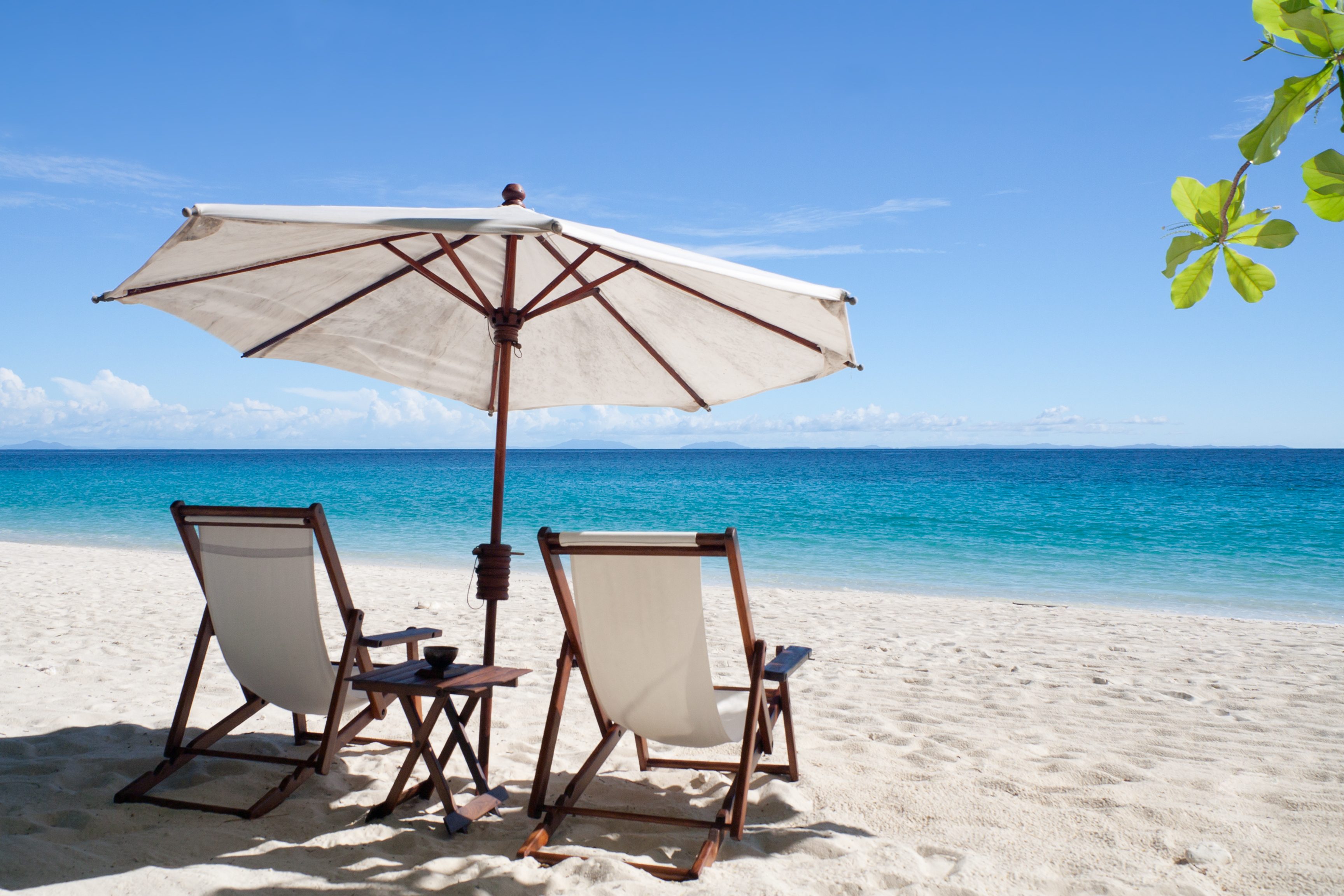 Two beach chairs under a large umbrella sit on white sand, overlooking calm blue ocean with clear skies and distant islands.