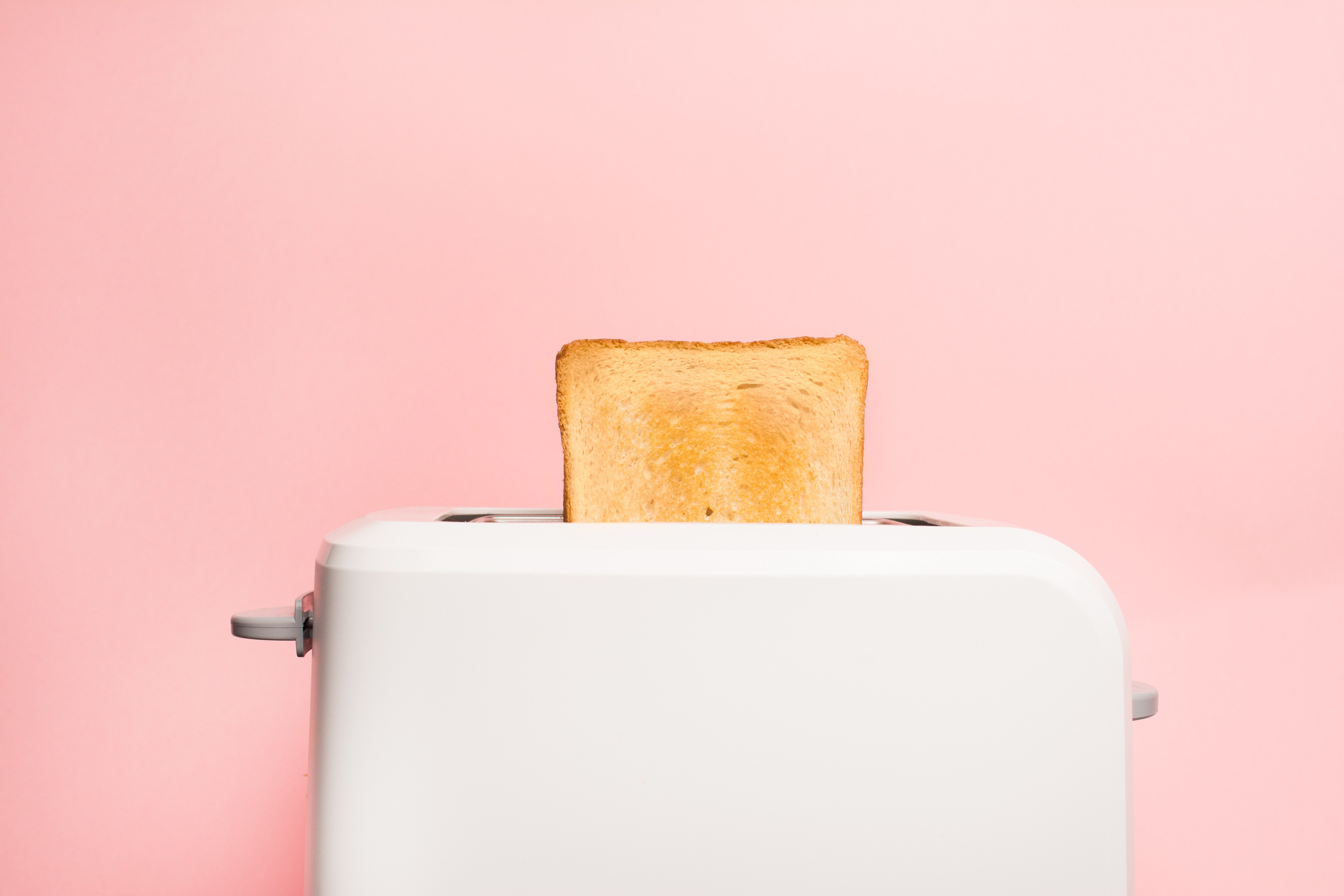 A white toaster ejects a slice of toasted bread against a pastel pink background.