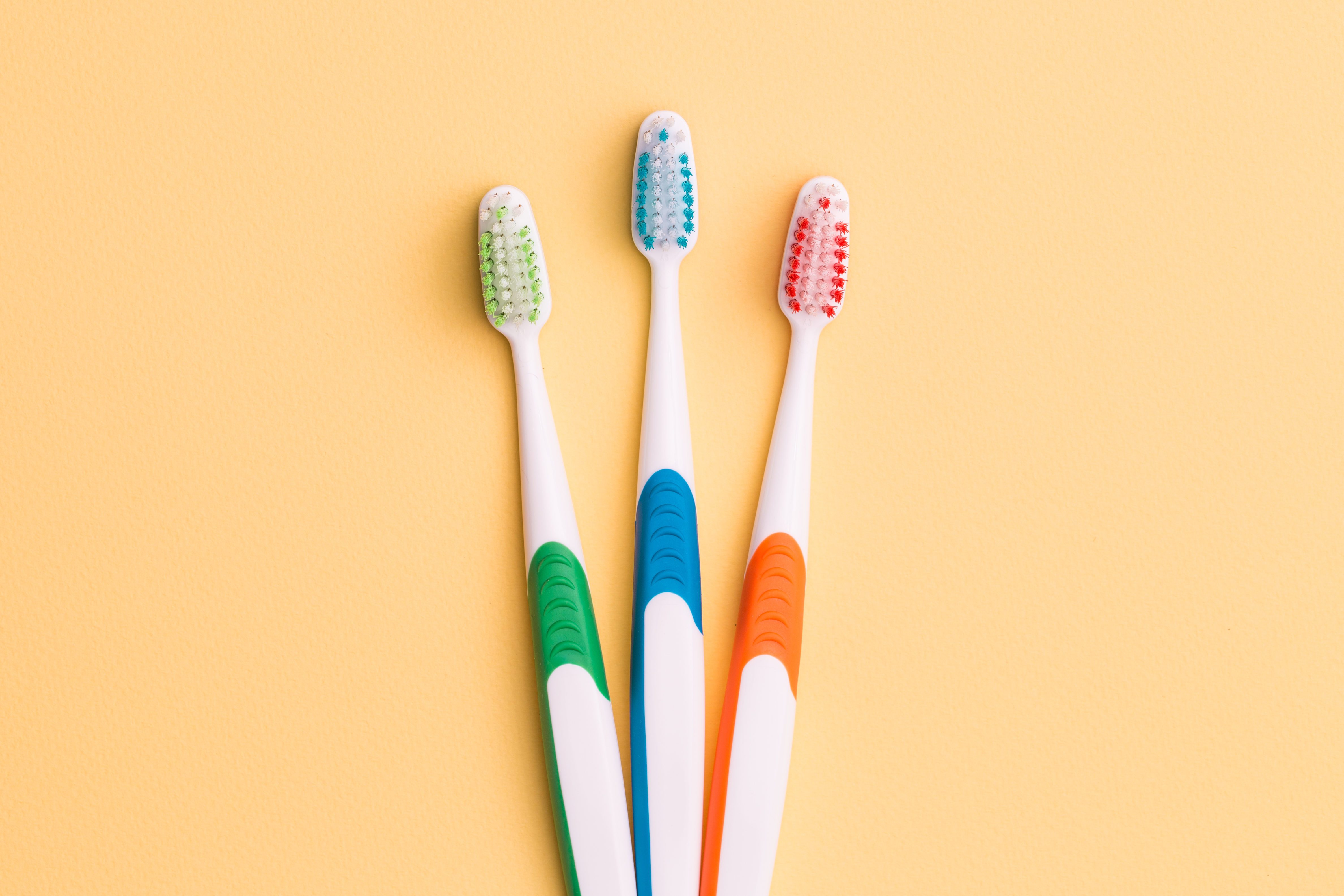 Three toothbrushes with colored bristles (green, blue, red) stand upright against a yellow background.