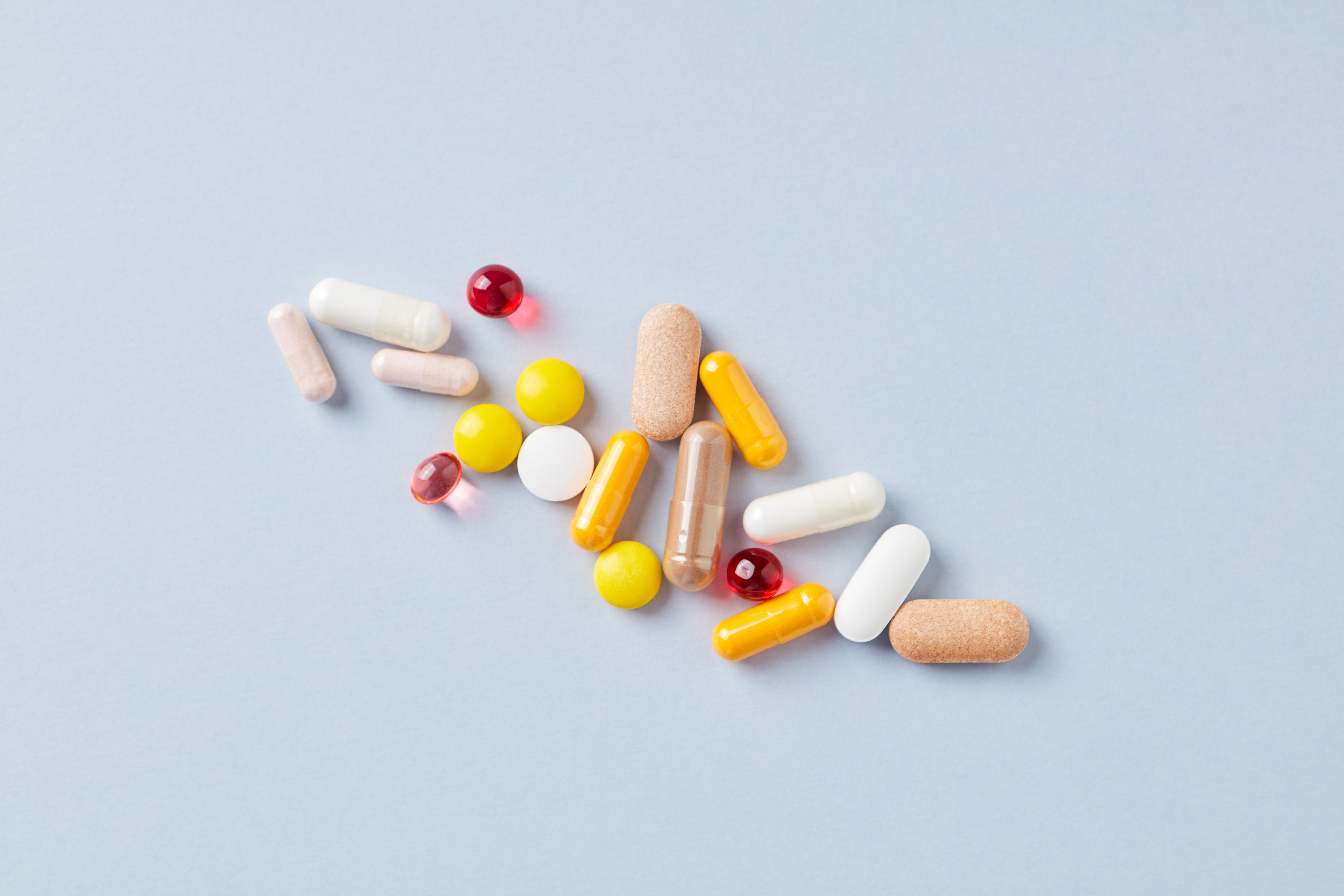 Assorted pills scattered on a light blue background, including capsules, tablets, and softgels in various colors.