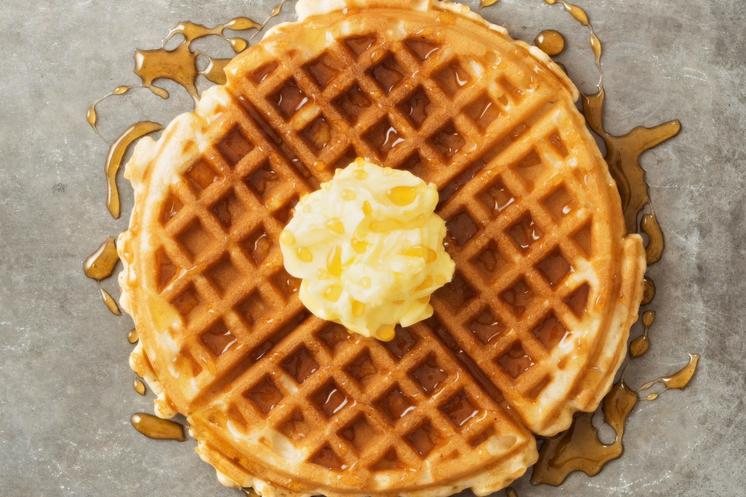 A waffle drizzled with syrup, topped with butter, sits on a gray surface.
