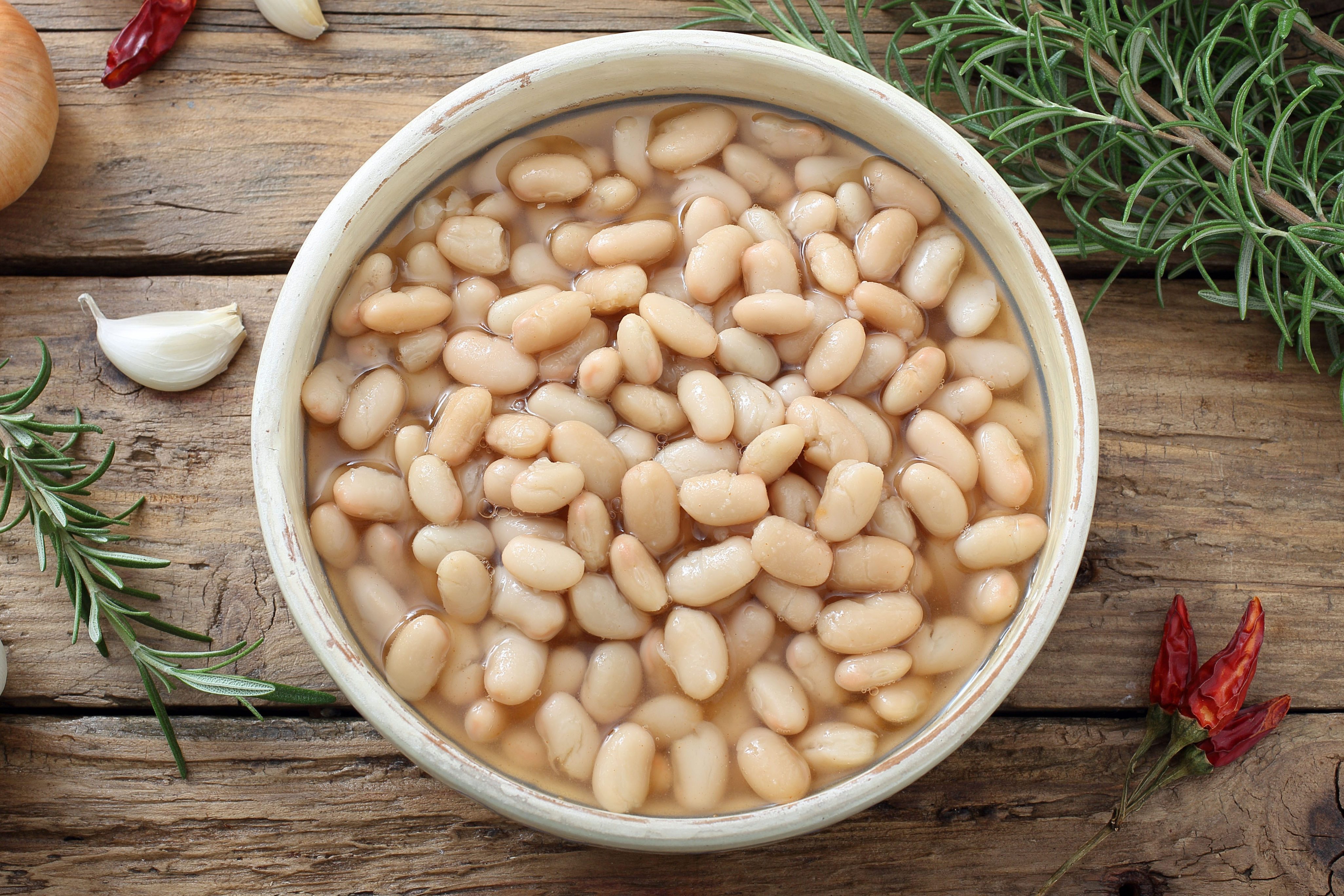A bowl of white beans sits on a wooden table surrounded by garlic cloves, rosemary sprigs, and dried chili peppers.