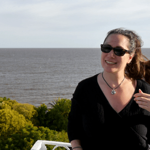 Person smiling, wearing sunglasses, on a balcony with lush greenery and ocean in the background.