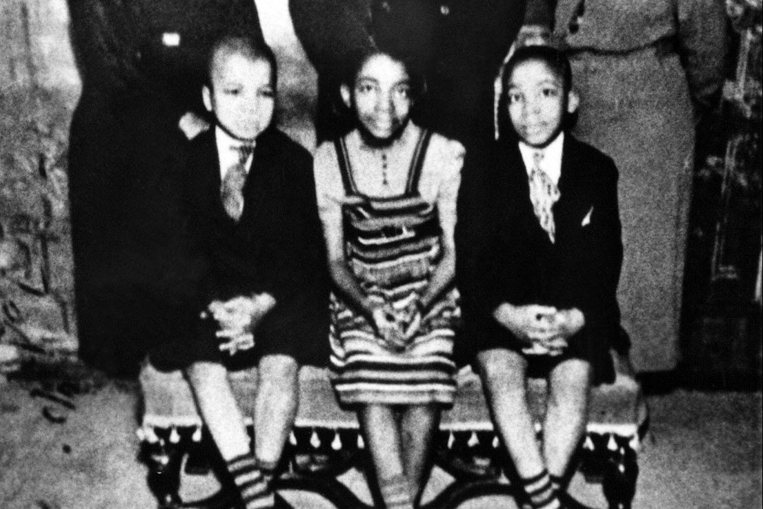 Three children sit on a bench, dressed in formal clothing, surrounded by standing adults in a vintage setting.