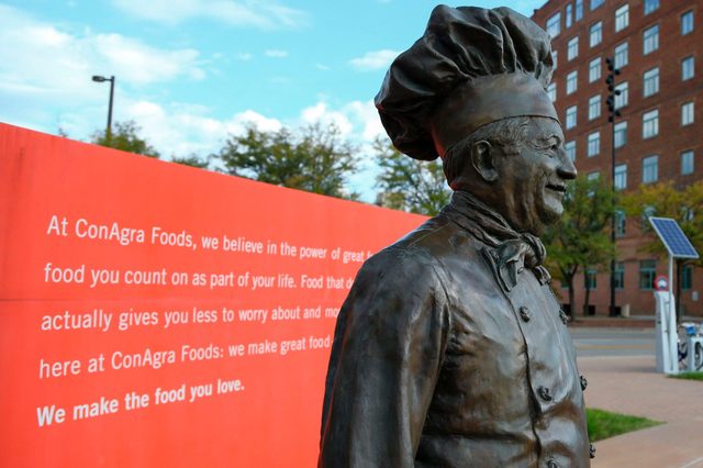 A bronze chef statue stands smiling, set against a red wall with text, in an urban outdoor area with trees and buildings.