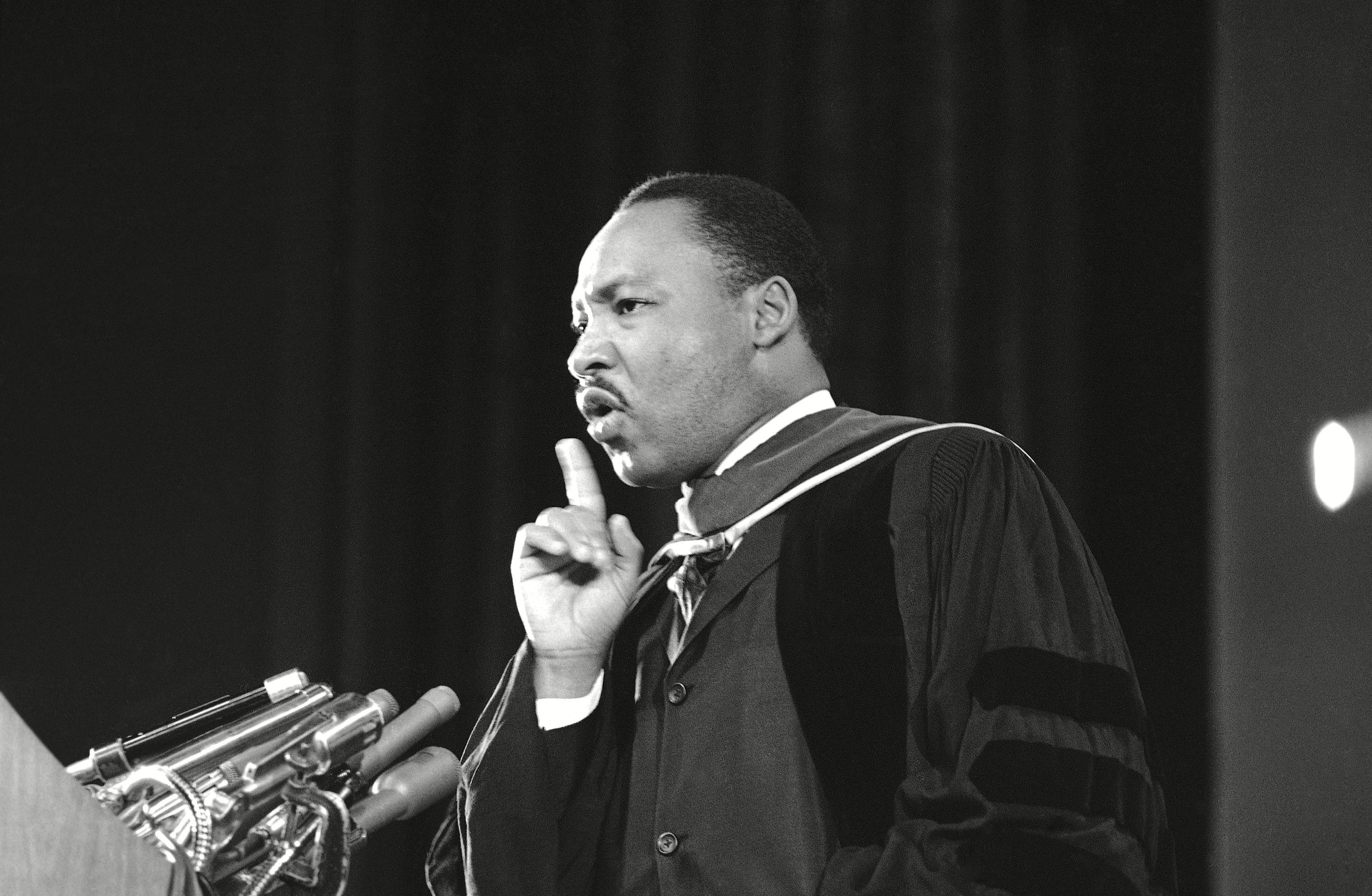 A speaker gestures emphatically while addressing an audience from behind a podium with multiple microphones in a formal indoor setting.
