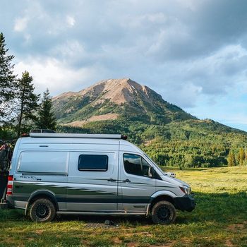 Van parked with bicycles attached, resting in a grassy field against a backdrop of towering, green-covered mountain under a cloudy sky.