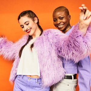 Two people smiling together, wearing colorful outfits, and dancing joyfully against an orange background.
