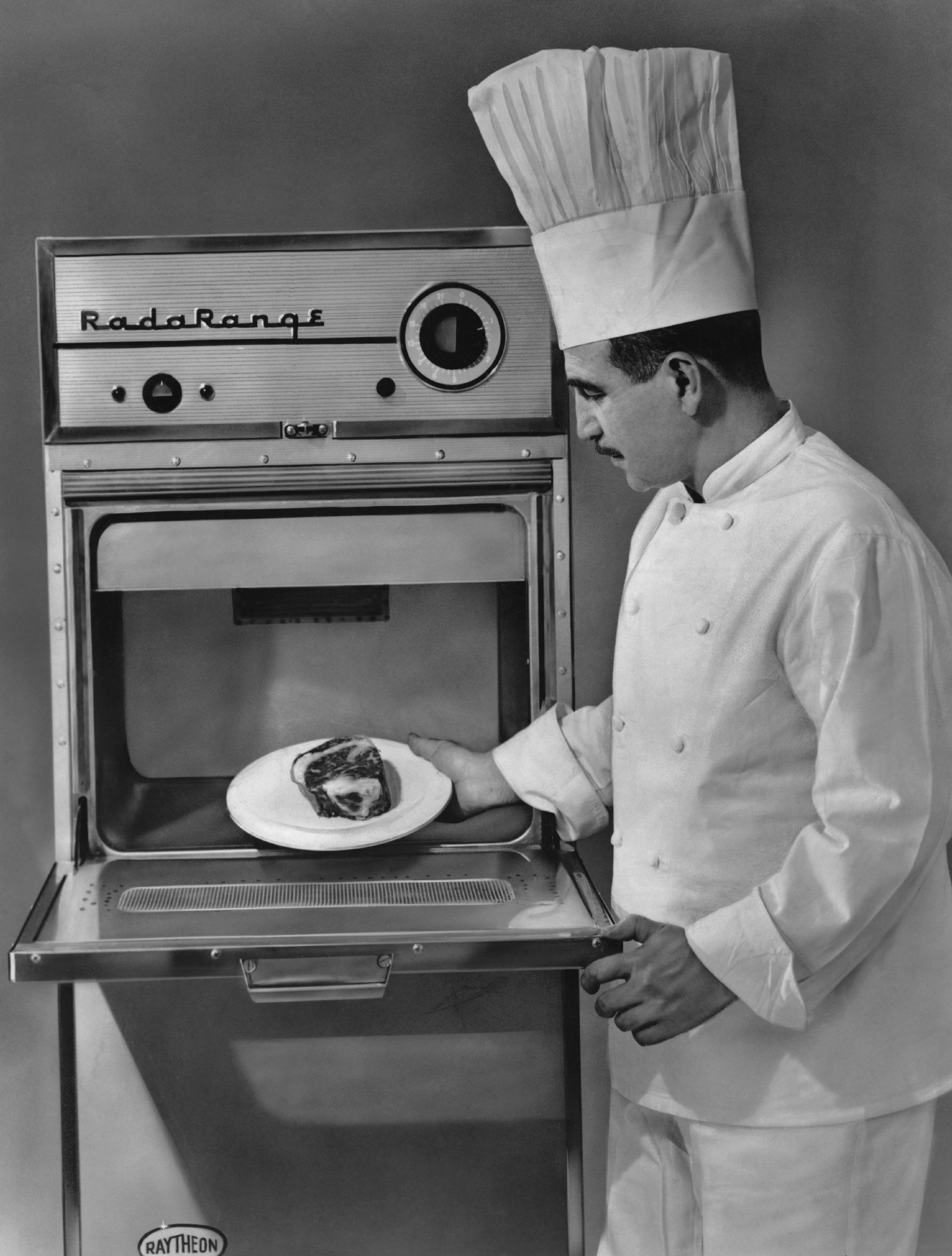 Chef places a plate with meat into an open "Rada Range" microwave; he's wearing a traditional white uniform and hat.