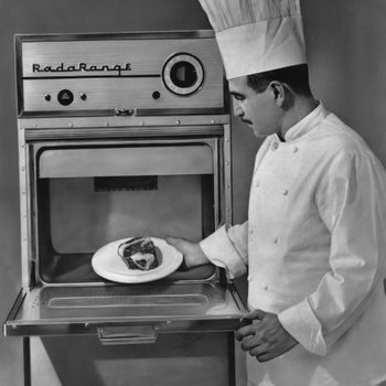Chef places a plate with meat into an open "Rada Range" microwave; he's wearing a traditional white uniform and hat.