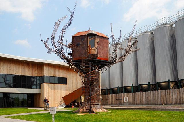 A whimsical treehouse with metal branches sits elevated beside a modern wood-paneled building and large silo structures, under a clear sky.