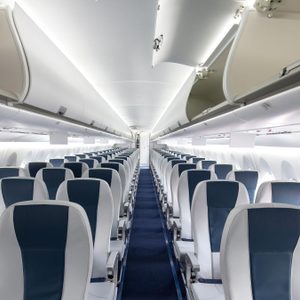 Empty airplane seats aligned, facing forward with overhead compartments open in a bright, modern aircraft interior.