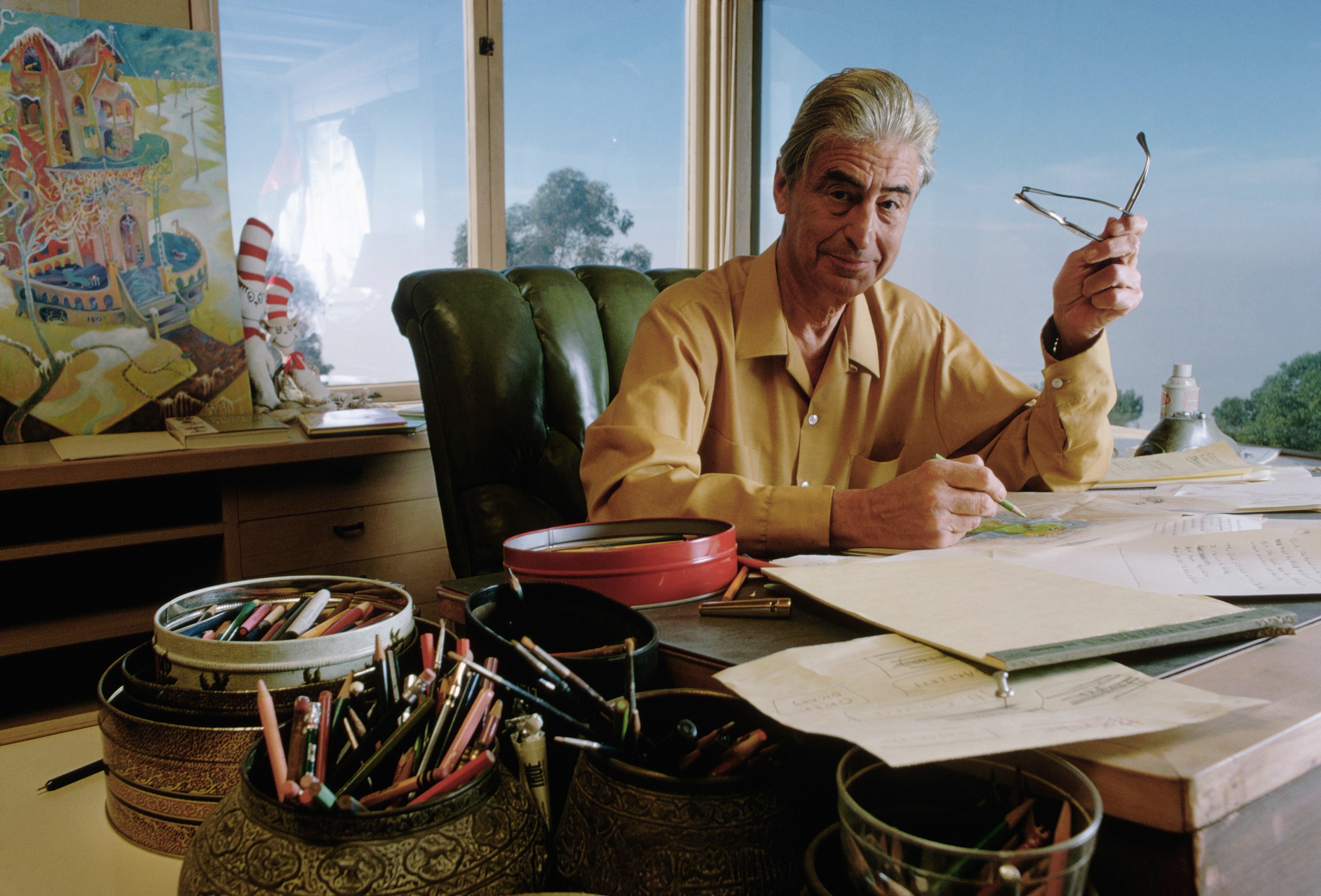 Man sitting, holding glasses, drawing on paper in a bright room. Art supplies and colorful painting nearby, large windows showing trees outside.