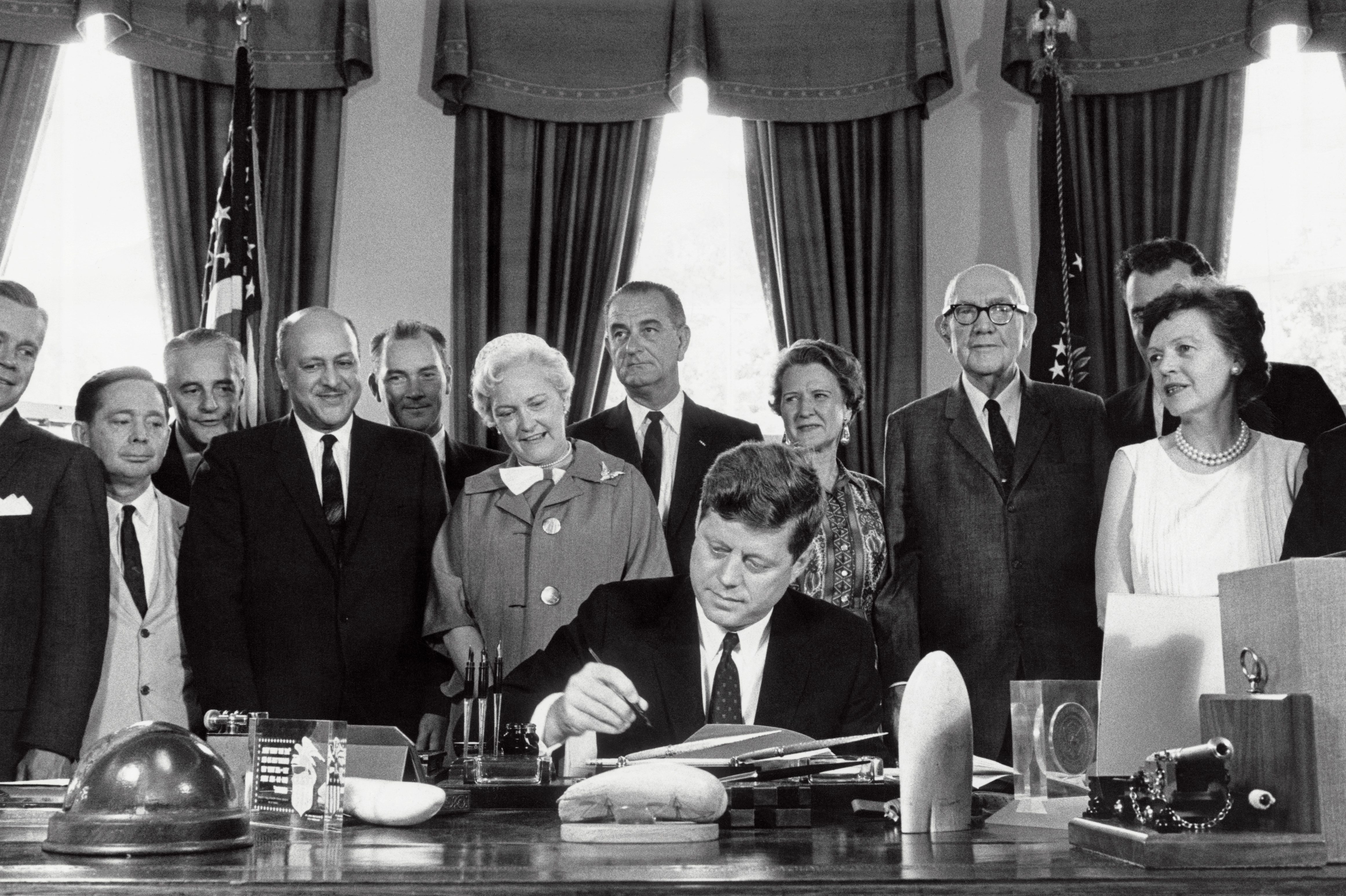A man signs a document at a desk, surrounded by smiling onlookers in suits and dresses, flags displayed in the curtained background.