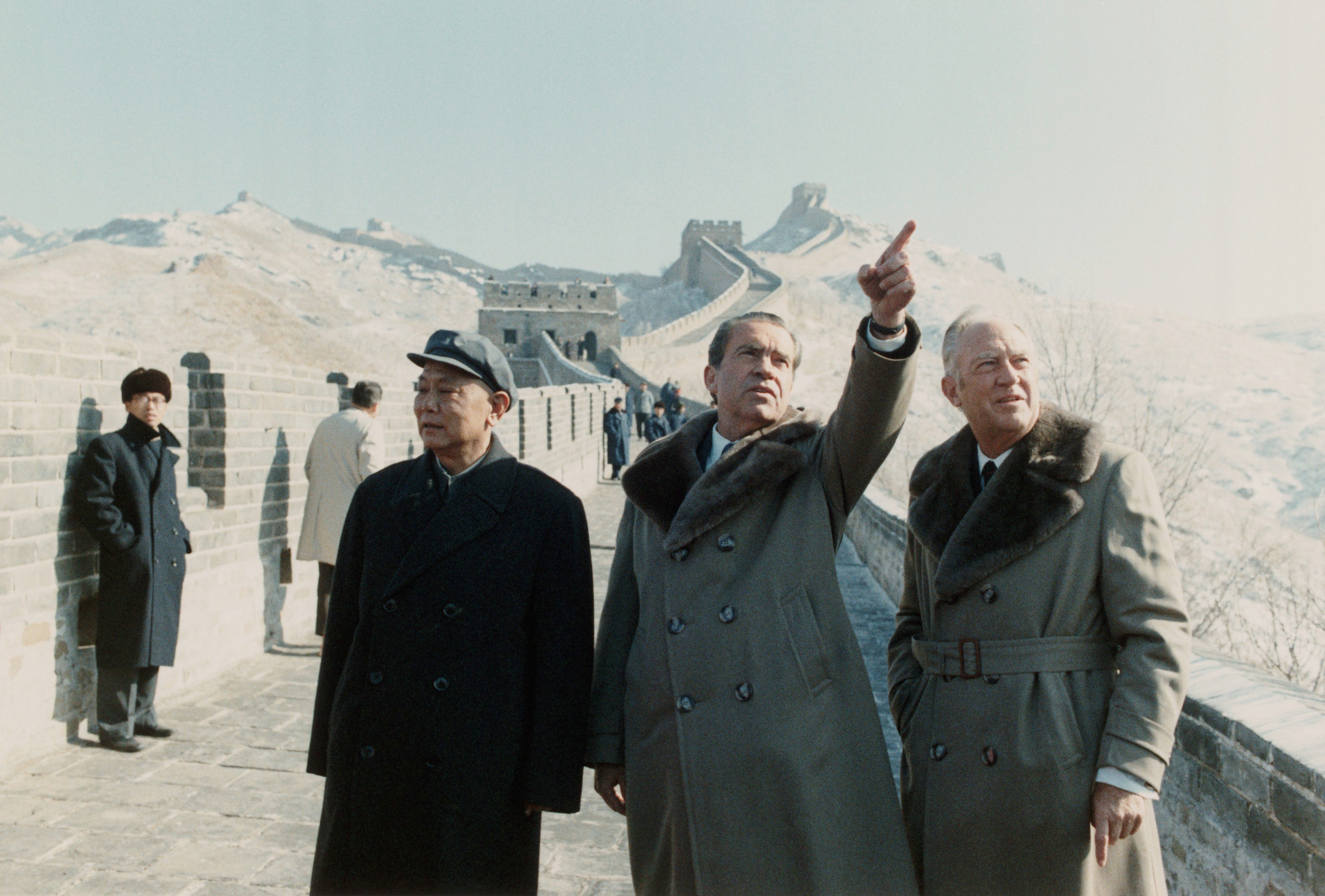 Three men in coats observe and discuss the Great Wall of China, with others walking nearby. Snowy mountains and stone walls form the background.
