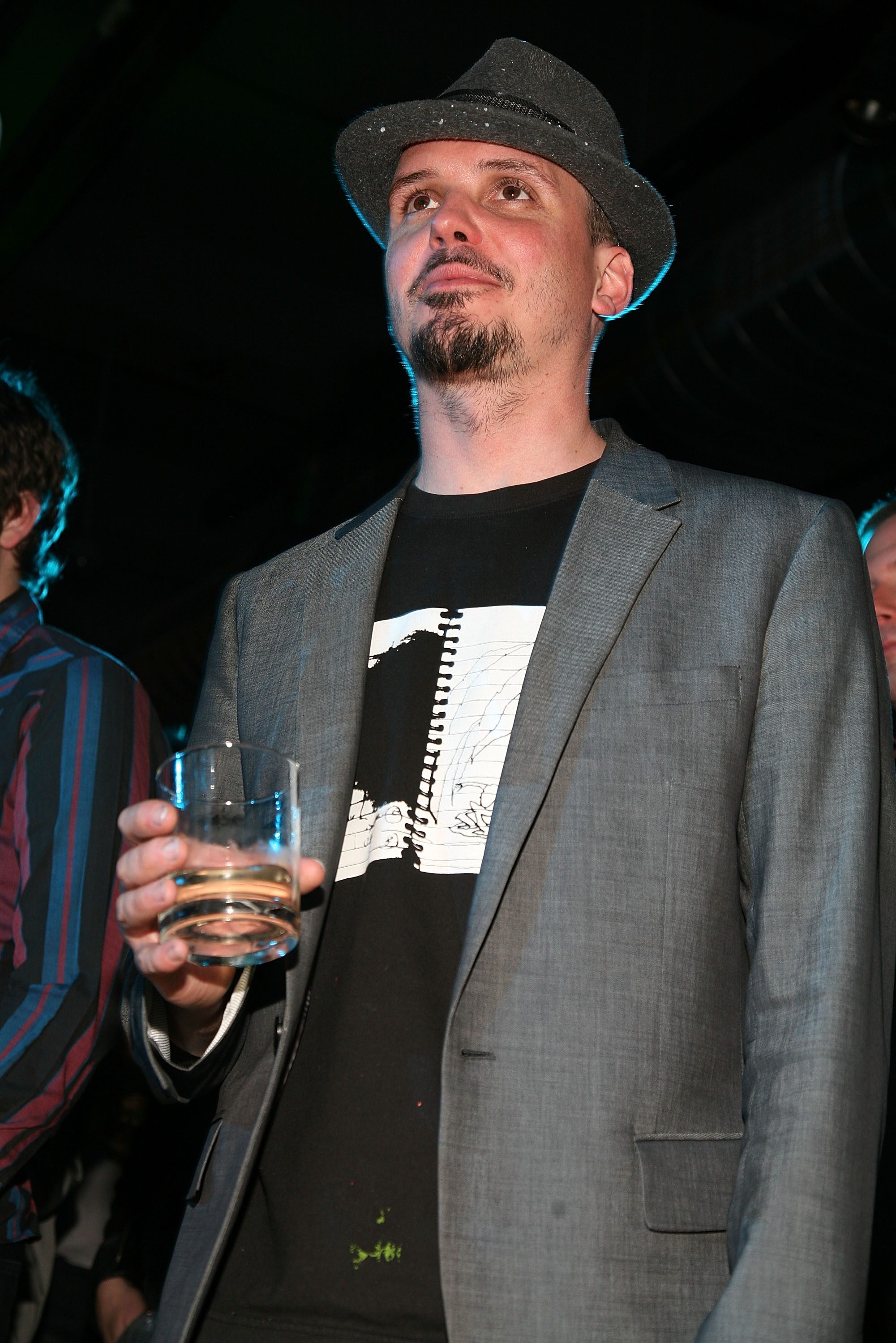 A man holding a drink stands in a dark environment, wearing a gray suit jacket, black shirt, and a gray fedora.