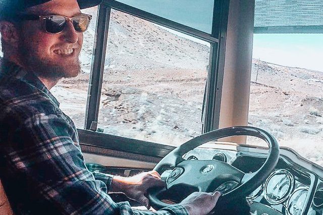 Man smiling, driving a bus through a desert landscape, wearing sunglasses and a plaid shirt.