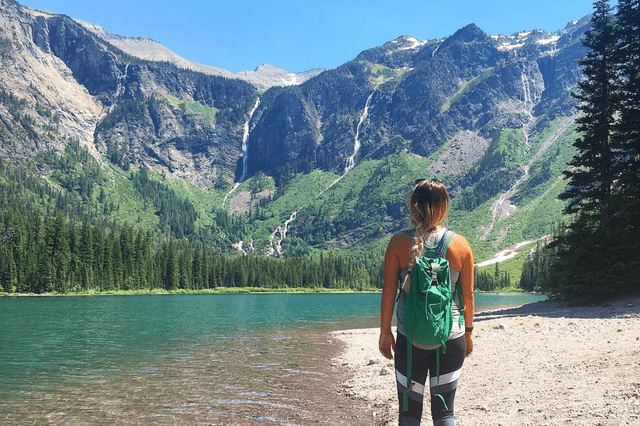 A person stands by a lake, overlooking mountains with waterfalls and trees under a clear blue sky.