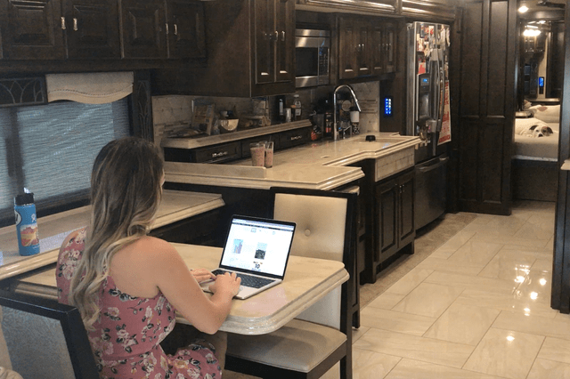 A woman types on a laptop at a small dining table inside a luxurious RV kitchen, with a dog visible on a bed.