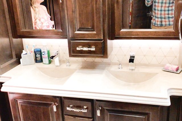 Double bathroom sinks sit beside each other; toothbrushes, soap, and various bottles are neatly arranged. A wooden cabinet with mirrors is above.