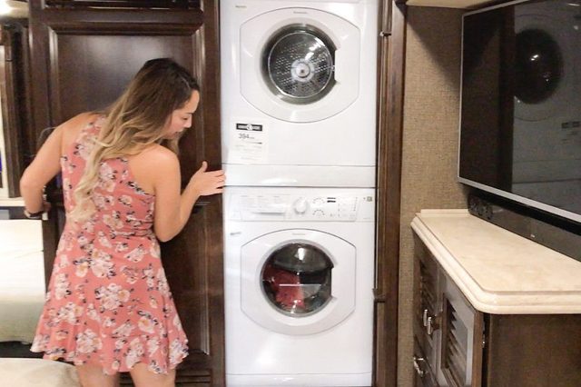 A woman inspects a stacked washing machine and dryer in a compact living space with wooden cabinets and a flat-screen TV nearby.