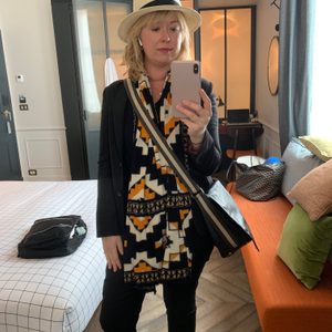 Person taking a mirror selfie, wearing a hat and patterned scarf, in a modern bedroom with a checkered bedspread and decorative pillows.