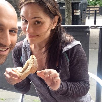 Two people smiling; one holds a sandwich outside a cafÃ©, with trees and a street visible in the background.