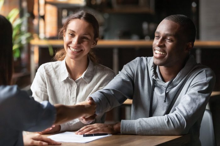 Happy young interracial couple shake hand of bank manager broker buy insurance services take mortgage loan, mixed ethnicity customers handshake agent make agreement financial business deal at meeting