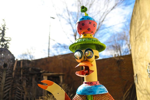 Whimsical sculpture stands colorful and wide-eyed outdoors, featuring a vibrant hat and spiral eyes, surrounded by bare trees and a wooden structure.